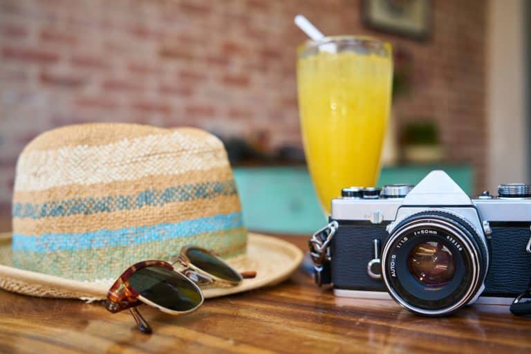 Photo d'un chapeau, à côté de lunettes de soleil, un appareil photo et un verre de jus, représentant un départ en vacances
